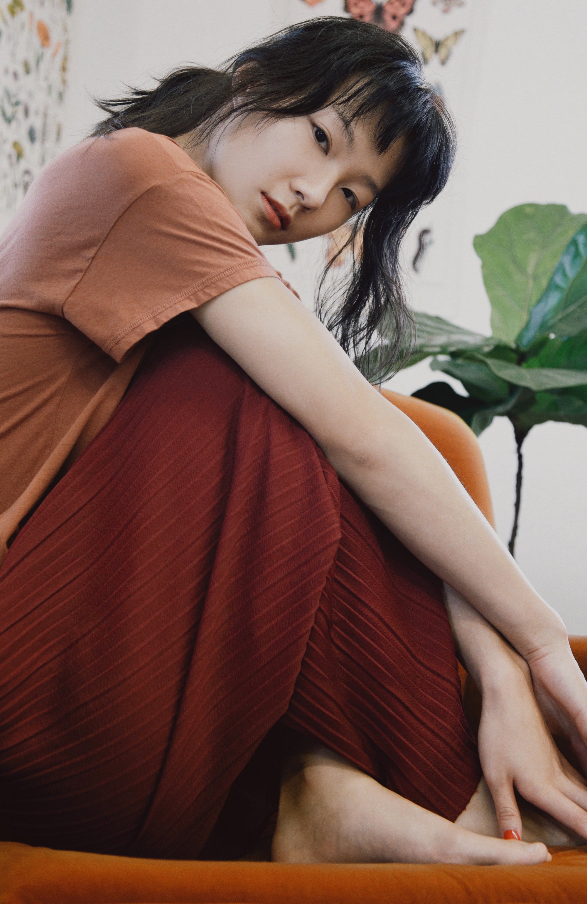 A woman sitting on an orange couch, wearing a rust-orange short-sleeve top and a dark red pleated skirt.